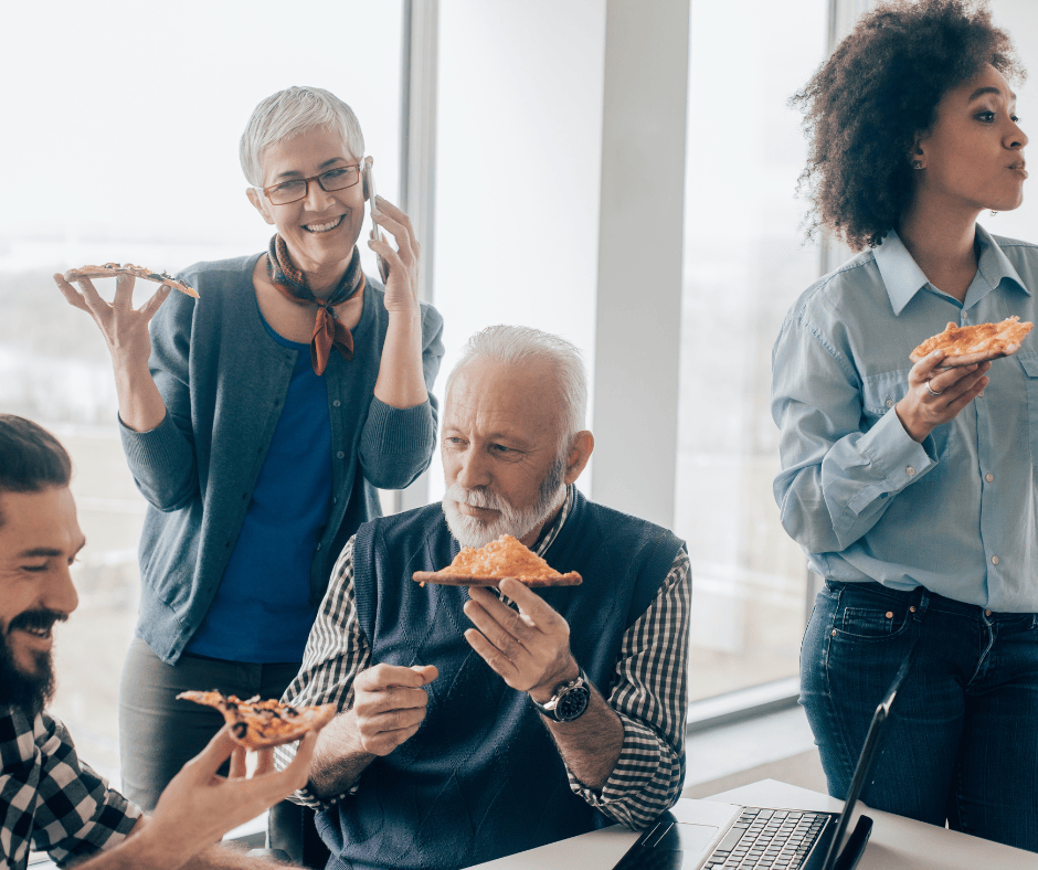 Eine ältere Frau und ein älterer Mann essen fröhlich mit einem jüngeren Mann und einer jungen Frau im Büro Pizza und kommunizieren miteinander, lachen oder arbeiten nebenbei.